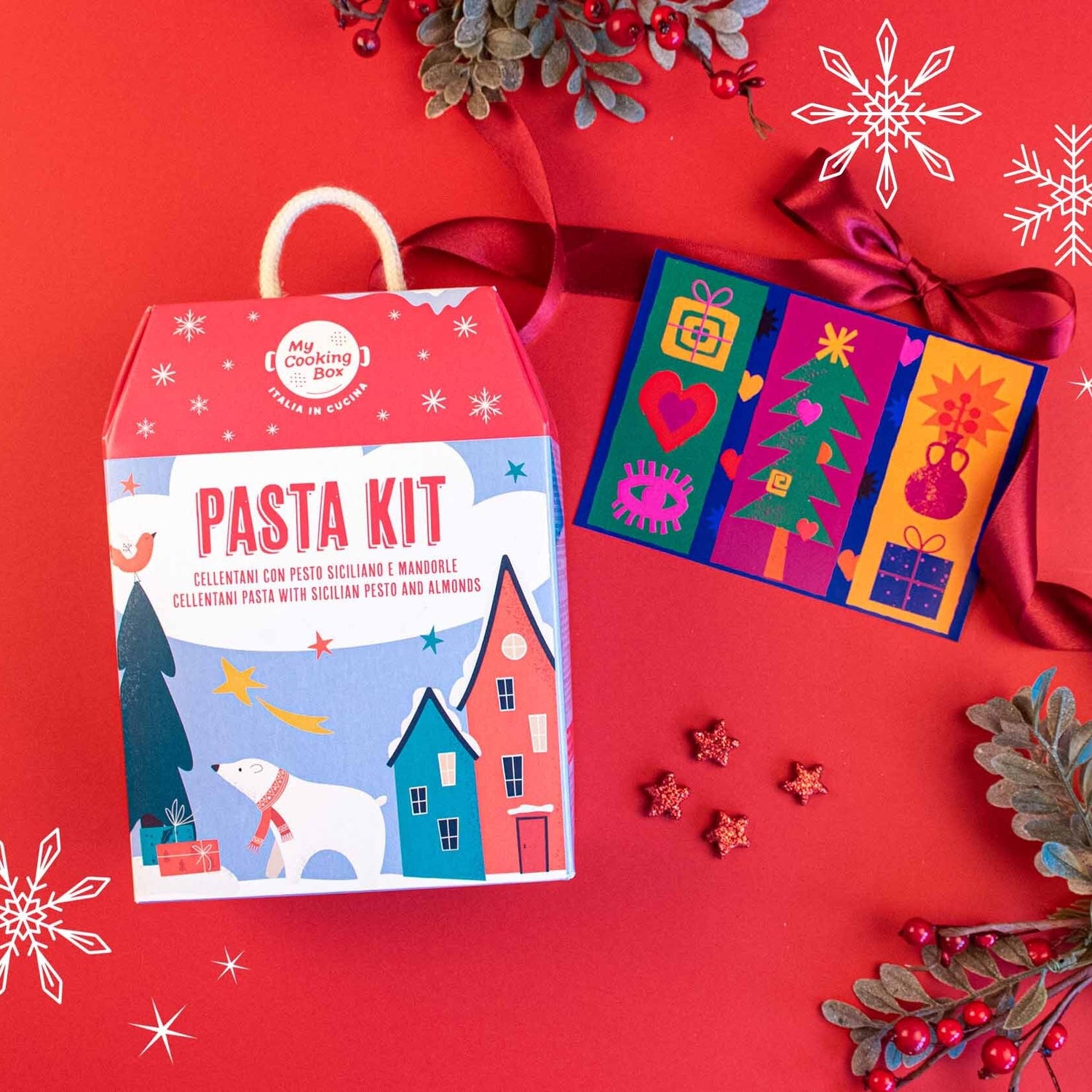 Pasta Kit Cellentani con pesto siciliano in una confezione natalizia e il biglietto solidale di Amref Health Africa su uno sfondo rosso e con elementi natalizi 
